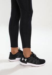 Svarta träningstights med texturerade paneler och en sidodragkedja, kombinerade med svarta och vita träningsskor med ovandel i mesh och logotyp.