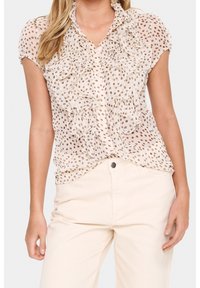 Blusa beige chiaro con volant e piccole macchie marroni, scollo dettagliato, maniche corte e bottoni sul davanti, abbinata a pantaloni di colore chiaro.