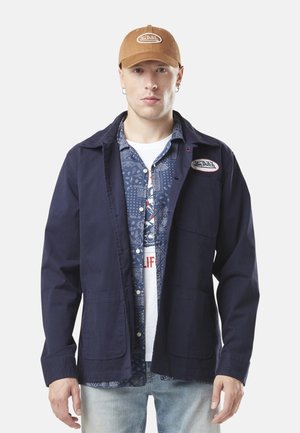 Homme portant une casquette beige Von Dutch, une veste bleu marine avec un patch, une chemise bleue à motifs, un t-shirt blanc à motif et un jean délavé, debout devant un fond blanc.