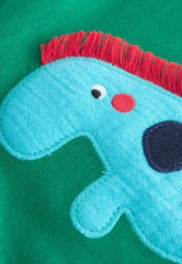 Aplique de tela azul con forma de dinosaurio, con flecos rojos, un punto negro y un círculo rojo sobre un fondo verde texturizado.