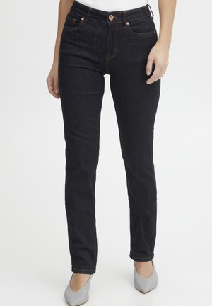 Kvinde iført mørkeblå slim-fit jeans med orange syninger og lys grå spidse hæle, der står foran en ensfarvet hvid baggrund.