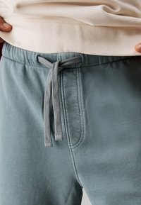 I pantaloni jogger in cotone blu chiaro presentano una vita elastica con un cordino grigio e tasche laterali. Il tessuto ha una consistenza morbida e liscia.