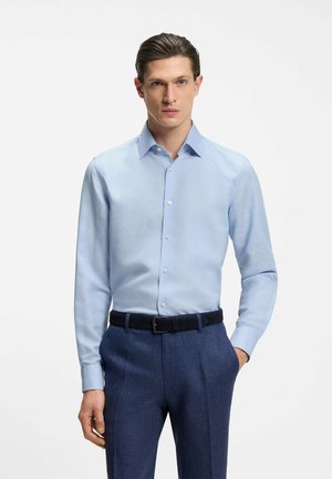 Mężczyzna ubrany w jasnoniebieską koszulę zapiętą na guziki, włożoną w ciemnoniebieskie spodnie z czarnym paskiem, stoi z jedną ręką w kieszeni.