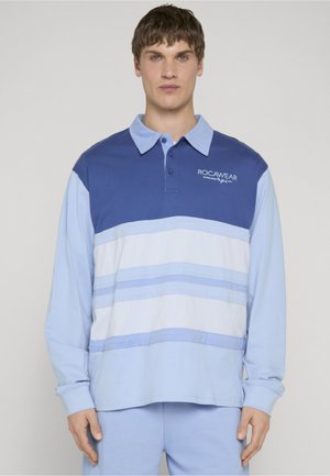 Giovane uomo che indossa una polo a maniche lunghe blu e bianca con righe orizzontali e il logo "ROCAWEAR" sul petto, in piedi davanti a uno sfondo semplice.