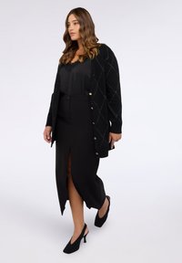 Cardigan negru cu model în romburi, nasturi și un top cu dantelă pe dedesubt. Asortat cu o fustă neagră cu o crapă frontală și tocuri negre.