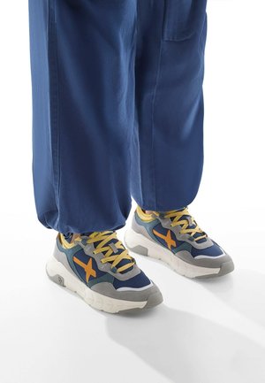 Sport sneakers with a gray upper, blue accents, and a prominent orange logo. Features yellow laces and a thick white sole.