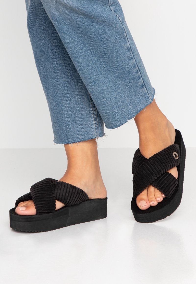 Pieds portant des sandales à plateforme noires avec des larges brides croisées en velours côtelé et un jean bleu court avec des ourlets effilochés.