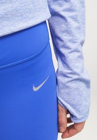 Leggings aderenti blu con una texture liscia e logo Nike argentato, abbinati a un top a maniche lunghe blu chiaro melangiato con polsini morbidi.