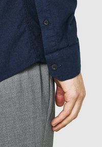 Camicia blu navy con finitura testurizzata. Presenta un orlo curvo e un polso con bottone. I pantaloni grigi sono parzialmente visibili sullo sfondo.