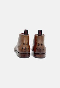 Botas de tobillo de cuero marrón con costuras decorativas, cortes en forma de diamante en el tacón, forro rojo y lengüetas en la parte posterior. Acabado pulido.