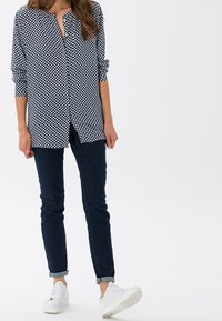 Blusa blu navy con motivo geometrico bianco, maniche lunghe e chiusura a bottoni. Abbinata a jeans scuri e sneaker bianche.