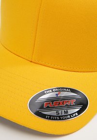 Kollane Flexfit nokamüts, millel on struktureeritud disain, tekstuuriga kangas, kõver visor ja silmapaistev must ja hõbedane logo silt.