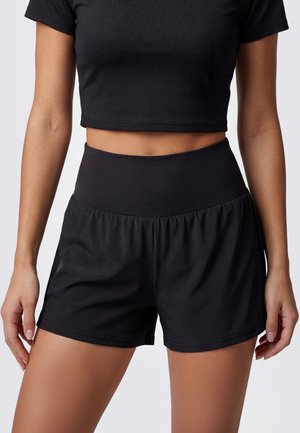 Donna che indossa pantaloncini sportivi neri a vita alta e un top corto nero a maniche corte su sfondo semplice.