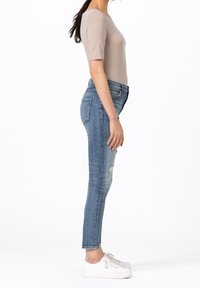 Kvinna i beige kortärmad topp, blå skinny jeans med lätt slitning, och vita sneakers som står i profil mot vit bakgrund.