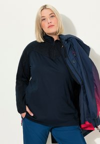 Camiseta de forro polar azul marino de manga larga con cuello con cremallera, que presenta un panel superior texturizado y mangas suaves. Combinada con pantalones azul claros livianos.