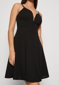 Kvinna som bär en ärmlös svart klänning med tunna axelband och en figurnära bodice med en djup V-ringning.