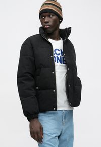 Junge Person mit einer gestreiften Mütze, schwarzer Steppjacke, weißem T-Shirt mit Grafik und hellblauen Jeans, die seitlich gegen einen schlichten Hintergrund schaut.