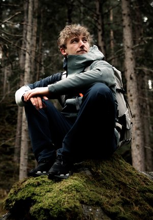 Young man in outdoor gear sitting on a moss-covered rock, resting in a dense forest with tall trees in the background.