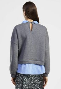 Donna vista di spalle che indossa un top grigio a maniche lunghe sopra una camicia colletto azzurra e una gonna con paillettes.