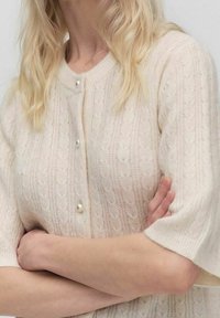 Světle béžový pletený kardigan s krátkými rukávy, s texturovaným vzorem a malými perlovými knoflíky vpředu. Měkký, hřejivý materiál.