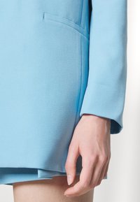 Giacca tailored azzurro chiaro con una superficie liscia, maniche lunghe e una tasca laterale discreta. L'orlo è leggermente svasato.