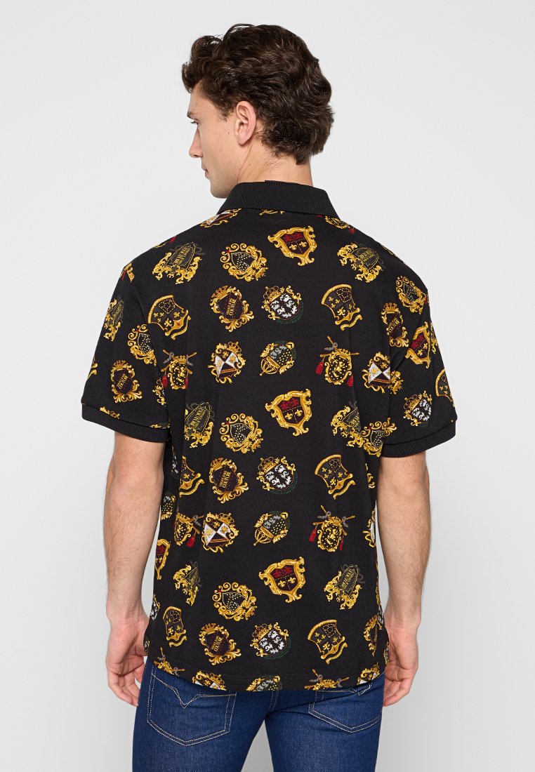 Mann, der ein schwarzes kurzärmliges Polohemd mit goldenen und roten, mittelalterlichen Wappenmustern trägt, kombiniert mit blauen Jeans, und von der Kamera abgewandt ist.