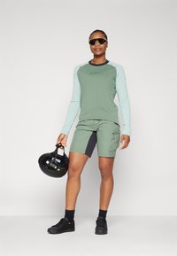 Lettvekts langermet skjorte i grønn med mintfargede ermer, kombinert med grønne shorts med mørkegrå aksenter. Svart hjelm holdes i hånden.