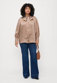 Brun mønstret bluse med puffede ærmer, bundet krave detalje, kombinert med utsvingte blå denimjeans og en liten brun veske.