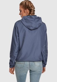 Könnyű sötétkék kabát kapucnival, rugalmas mandzsettákkal és sima felülettel, világos farmernadrág fölött viselve. Közeli nézet hátulról.