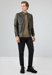 Bruine leren jas met ribbelontwerp op de schouders, ritsen en twee voorkruiken. Gekozen boven een groene shirt en gecombineerd met zwarte jeans en sneakers.