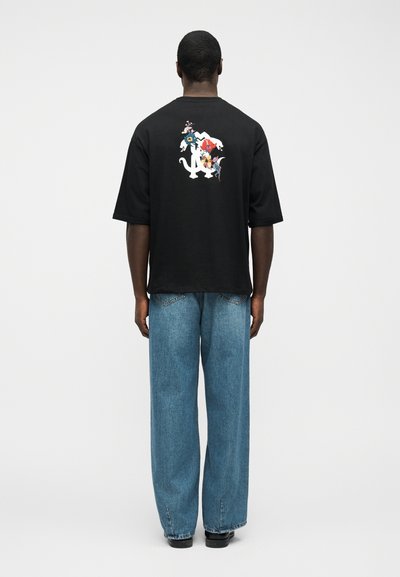 Άντρας που φοράει μαύρο μεγάλο t-shirt με φλοράλ και ζωακό σχέδιο στην πλάτη, συνδυασμένο με φαρδιά μπλε τζιν και μαύρα παπούτσια, στέκεται γυρισμένος προς τα πίσω.