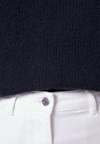 Gros plan d'un pull noir tricoté au-dessus de la taille d'un jean en denim blanc avec une fermeture par bouton argenté.
