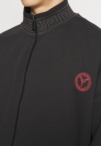 Schwarze Zip-Up-Jacke mit einem strukturierten Kragen im geometrischen Muster und einem roten gestickten Logo auf der Brust. Weiche Stofftextur.
