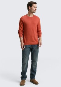 Mann, der seitlich steht, mit einem rostfarbenen Langarmpullover, blauen Jeans und braunen Wildlederstiefeln vor einem hellen Hintergrund.