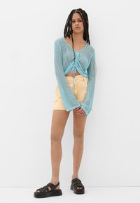 Helles blauer Strickpullover mit langen Ärmeln und Kordelzugdetail, kombiniert mit gelben Shorts und schwarzen Plateausandalen.