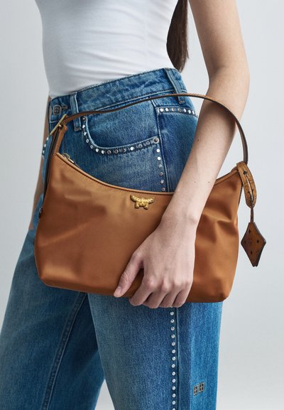 Kvinna i nitar jeans i blått och vit topp som håller en brun handväska med guldemblem och läderrem mot en enfärgad bakgrund.