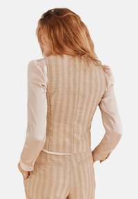 Beige linnen vest met verticale strepen, gecombineerd met een licht mouwloos topje. Gelaagd stof, getailleerde pasvorm, en twee achterzakken zichtbaar.