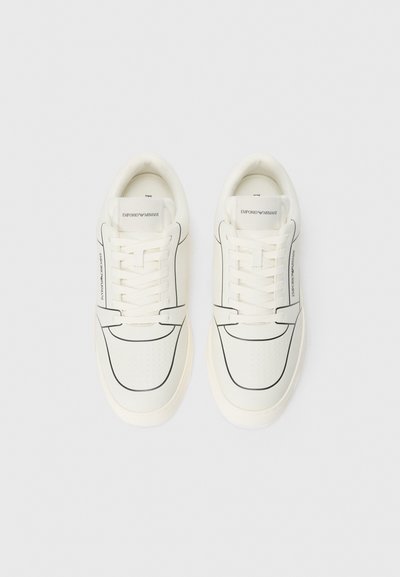 Baskets blanches avec une texture perforée, des détails en contour noir, des lacets plats et un logo sur la languette, fabriquées en matériau synthétique lisse.