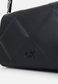 Svart quiltad handväska i läder. Har en texturerad yta och ett präglat "CK"-logotyp. Kedjesträngen synlig på ena sidan.