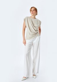 Beige draperige top met korte mouwen, gecombineerd met witte wijde broek en puntige witte hakken met decoratieve accenten.