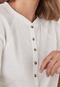 Gros plan sur une personne portant une chemise à manches longues texturée blanche avec cinq boutons bruns, la main reposant à proximité.