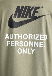 Olivinės spalvos marškinėliai su dideliu juodu Nike logotipu ir balta užrašu "Tik įgalioti asmenys." Pagaminti iš glotnaus, lengvo audinio.