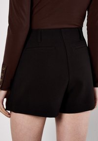 Svarta skräddarsydda shorts med en slät yta, med två baktoi fickor och en ren skärning. Kombinerat med en figurnära brun långärmad topp.