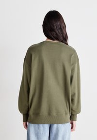 Olajzöld, oversized mikulás pulóver, raglán ujakkal és bordás mandzsettákkal és aljjal. Puha tapintás, minimalista dizájn, világoskék farmerrel viselve.