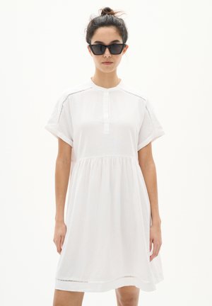 Vestito bianco a maniche corte con colletto bottonato, vita arricciata e dettagli in pizzo lungo le cuciture, abbinato a occhiali da sole neri con forma angolare.