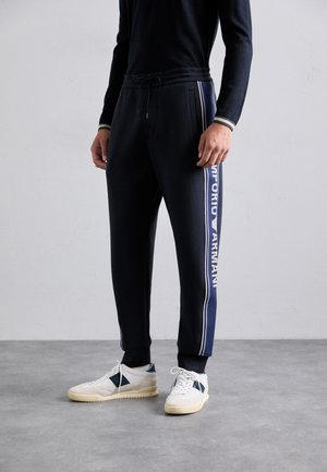 Bărbat îmbrăcat în pantaloni de trening tapered negri cu dungi albastre pe lateral și text alb, purtând adidași albi și negri, stând pe un podea gri.