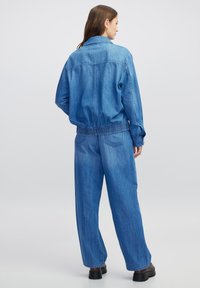 Vrouw die met de rug naar de camera staat in een blauwe spijkerjas en wijde spijkerbroek met zwarte schoenen op een effen lichte achtergrond.