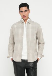Joven con una camisa de cuadros beige con botones sobre un suéter blanco de media cremallera y pantalones de cuero negros, de pie con las manos en los bolsillos.