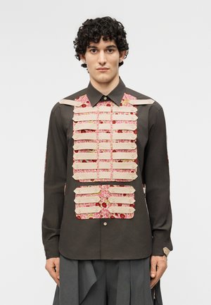 Homme portant une chemise sombre avec des panneaux de tissu floral superposés maintenus par des sangles beige au niveau de la poitrine et du ventre, debout devant un fond uni.