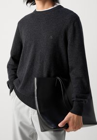 Dunkelgrauer Strickpullover mit gerippten Bündchen und Saum, kombiniert mit einer glänzenden schwarzen Tragetasche mit flacher Silhouette und langen Henkeln.